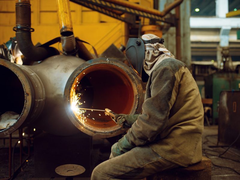 Welder in mask works with big metal pipe on factory, welding skill. Metalworking industry, industrial manufacturing of steel products